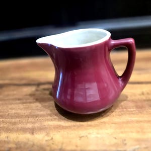 May include: A small, burgundy-colored ceramic creamer with a white interior. The creamer has a handle and a spout for easy pouring. It sits on a wooden surface.
