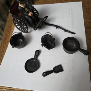 May include: A collection of miniature black metal toys, including a rickshaw with a seated figure, buckets, a frying pan, a ladle, and a small shovel. The toys are arranged on a white surface.