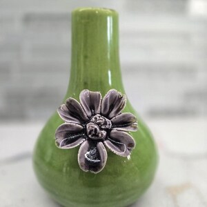 May include: A small, green ceramic vase with a bulbous body and a narrow neck. A dark gray flower with detailed petals is affixed to the front. The vase is approximately 6 inches tall and is suitable for small floral arrangements.