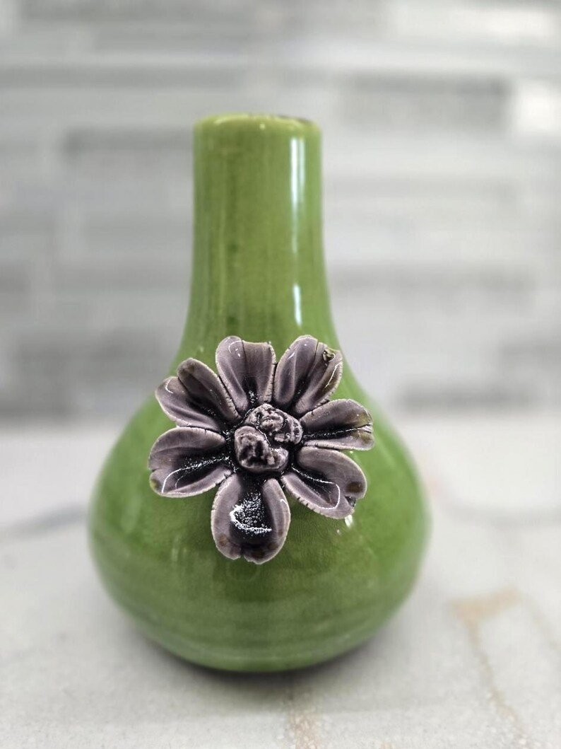 May include: A small, green ceramic vase with a bulbous body and a narrow neck. A dark gray flower with detailed petals is affixed to the front. The vase is approximately 6 inches tall and is suitable for small floral arrangements.