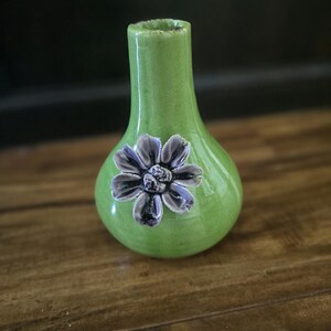May include: A small, green ceramic vase with a bulbous body and a narrow neck. A detailed, gray and purple flower is affixed to the front. The vase is displayed on a wooden surface.