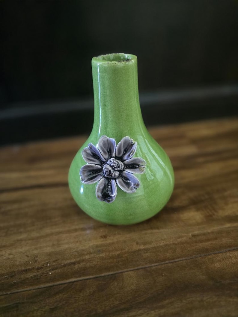 May include: A small, green ceramic vase with a bulbous body and a narrow neck. A detailed, gray and purple flower is affixed to the front. The vase is displayed on a wooden surface.