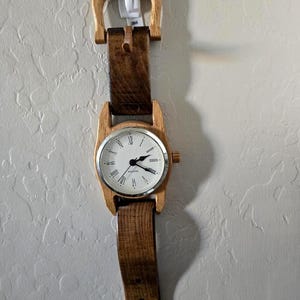 May include: A unique wall clock designed to resemble a wristwatch. The clock features a light-colored wooden frame and a brown leather strap. The clock face is white with black hands and Roman numeral hour markers. The wooden buckle is at the top.