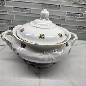 May include: A white porcelain tureen with a matching lid, featuring delicate floral patterns in pink and green, gold-trimmed edges, two side handles, and a decorative finial on the lid, all set against a smooth, rounded, embossed base.
