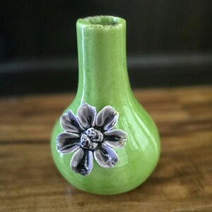 May include: A small, green ceramic vase with a bulbous body and a narrow neck. A detailed, gray and purple flower is affixed to the front. The vase is approximately 6 inches tall and is suitable for displaying small floral arrangements.