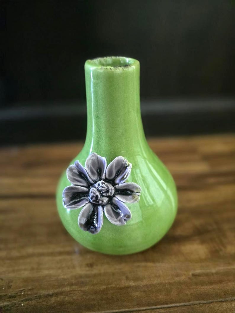 May include: A small, green ceramic vase with a bulbous body and a narrow neck. A detailed, gray and purple flower is affixed to the front. The vase is approximately 6 inches tall and is suitable for displaying small floral arrangements.