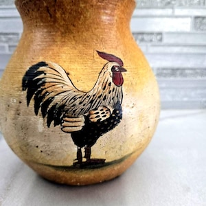May include: A handcrafted, rustic pottery vase with a rooster illustration. The vase has a wide, rounded body and a slightly flared rim. The rooster is depicted in black, white, and red, adding a touch of country charm to the decorative piece.