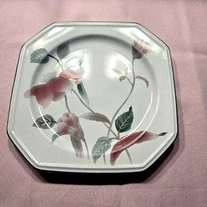 May include: An octagonal white plate with a black rim, featuring a floral design. The flowers are pink and the leaves are green and brown. The plate is on a pink surface.