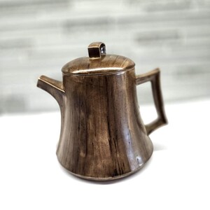 May include: A brown ceramic teapot with a lid and handle. The teapot has a textured surface with vertical lines, giving it a wood-like appearance. The spout is on the left side, and the handle is on the right.
