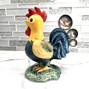 May include: A ceramic rooster figurine with a red comb, yellow head, and blue body. The rooster stands on a green base and has silver metal attachments on its back. The figurine is a decorative item.