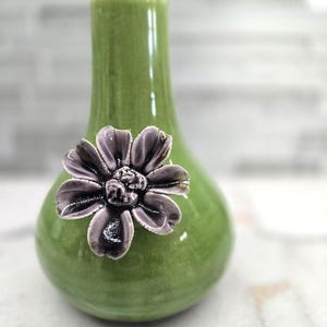 May include: A small, green ceramic vase with a glossy finish. A detailed, dark purple flower is affixed to the front of the vase. The vase has a rounded body and a narrow neck, suitable for displaying small floral arrangements.