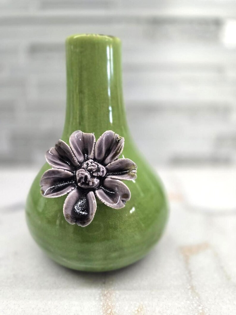 May include: A small, green ceramic vase with a glossy finish. A detailed, dark purple flower is affixed to the front of the vase. The vase has a rounded body and a narrow neck, suitable for displaying small floral arrangements.