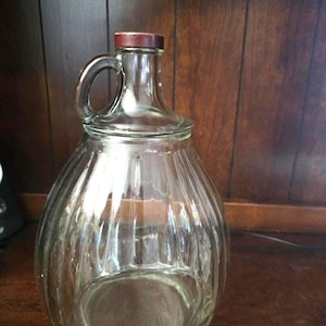May include: A clear glass jug with a rounded, ribbed body and a narrow neck featuring a single handle. The jug is topped with a small, circular, dark reddish-brown cap. The glass is transparent, showcasing vertical ridges that run along the sides.