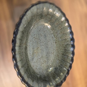 Puede incluir: Una fuente ovalada de cerámica con un interior verde apagado moteado y un borde festoneado marrón oscuro. El plato tiene un aspecto rústico y hecho a mano, adecuado para servir aperitivos o platos pequeños.