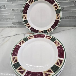May include: A stack of white ceramic plates featuring a wide rim decorated with a repeating geometric pattern. The design consists of alternating dark green and deep burgundy segments separated by tan triangles, all outlined in thin dark green lines.