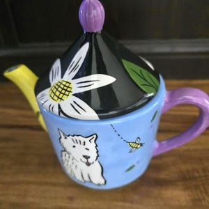 May include: A whimsical teapot with a black lid adorned with a white daisy and green leaf design. The teapot has a light blue body with a white dog illustration and a yellow spout. The handle is purple.