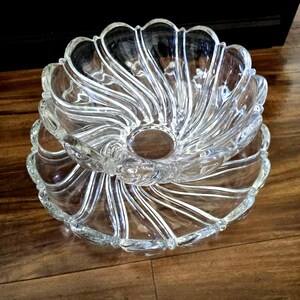 May include: Clear glass serving dish set. The set includes a large, decorative bowl with a scalloped edge, resting on a matching plate. The bowl and plate have a spiral design radiating from the center. The set is made of clear glass.
