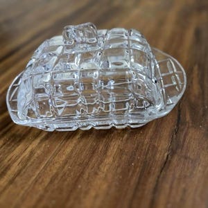 May include: Clear glass butter dish with a lid. The dish has a rectangular shape with a grid pattern design. The lid has a small handle on top. The butter dish is sitting on a wooden surface.