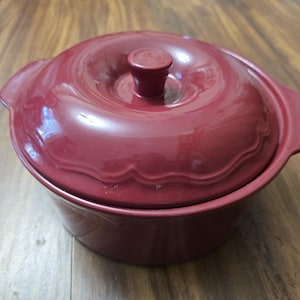 May include: A burgundy ceramic casserole dish with a matching lid. The lid has a central knob and decorative scalloped edges. The dish has two side handles and is set on a wooden surface.