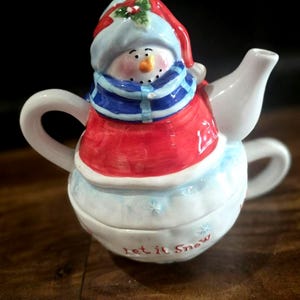 May include: A ceramic snowman teapot with a red coat, blue scarf, and a blue and white hat. The teapot has a white base with the words "Let it Snow" and snowflake details. The teapot has a handle and a spout.
