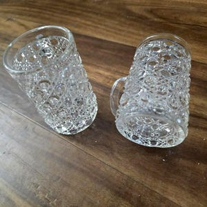 May include: Two clear, patterned glass mugs with handles. The mugs have a textured, cut-glass design. They are resting on a wooden surface. The mugs are likely for drinking beverages.