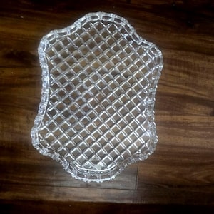 May include: A clear glass serving tray with a scalloped edge and a raised diamond grid pattern across the surface. The tray features a reflective, transparent finish with a structured geometric design that creates a textured appearance.