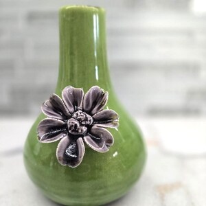 May include: A small, green ceramic vase with a bulbous base and a narrow neck. A decorative, dark purple flower is affixed to the front. The vase has a glossy finish and is likely for displaying small flowers or as a decorative accent.