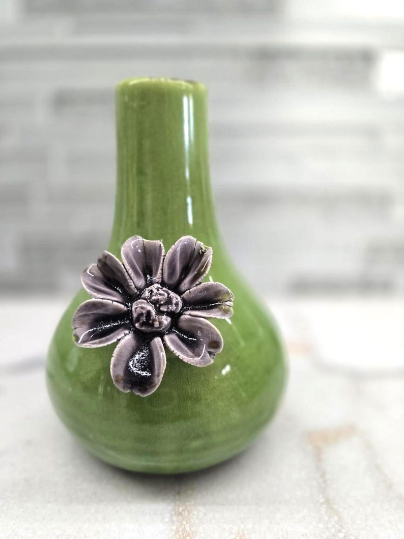 May include: A small, green ceramic vase with a bulbous base and a narrow neck. A decorative, dark purple flower is affixed to the front. The vase has a glossy finish and is likely for displaying small flowers or as a decorative accent.