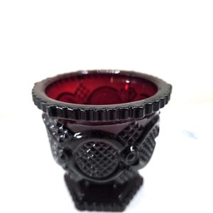 May include: A ruby red glass vase with a detailed, textured design. The vase has a wide, scalloped rim and a geometric pattern on the body. The base is a dark, solid color.