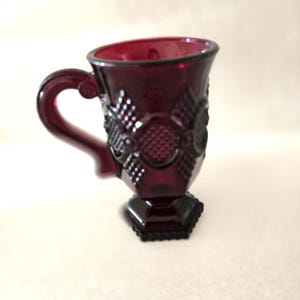 May include: A ruby red glass cup with a handle. The cup has a textured, patterned design and sits on a hexagonal base. The handle is curved and attached to the side of the cup. The cup is likely vintage or antique.