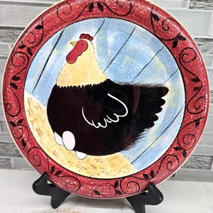 May include: A decorative ceramic plate featuring a hen illustration. The plate has a red border with a black scroll design. The center of the plate has a blue background with a hen in black, white, and yellow.