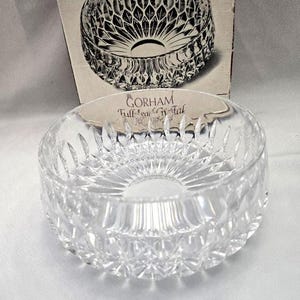 May include: A clear, round crystal bowl with a detailed, cut-glass design. The bowl is sitting in front of its original box, which features a graphic of the bowl. The text "GORHAM Full-lead Crystal" is visible on the bowl.