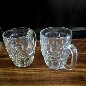 May include: Two clear glass beer mugs with textured sides and curved handles. The mugs are sitting on a wooden surface. The mugs are empty and ready to be filled with a cold beverage.