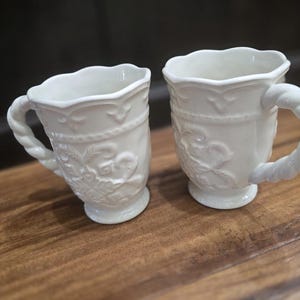 May include: Two white ceramic mugs with ornate floral and rope-like handle designs. The mugs have a scalloped rim and are set on a wooden surface. The mugs are likely for coffee or tea.