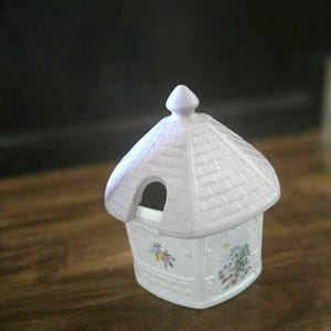 May include: A white ceramic trinket box shaped like a small house with a light purple roof and floral accents. The house has a small opening and a decorative finial on top. The box is set on a wooden surface.