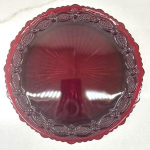 May include: A round, deep red glass plate featuring a scalloped edge and a decorative border with a repeating lattice pattern. The center of the plate displays a sunburst design radiating from the middle, with a smooth, glossy finish throughout.