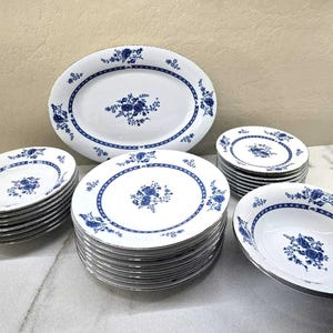 May include: A collection of white ceramic dinnerware featuring a blue floral pattern and a blue geometric border. The set includes one large oval platter, a stack of round dinner plates, a stack of smaller round plates, and a single round bowl.