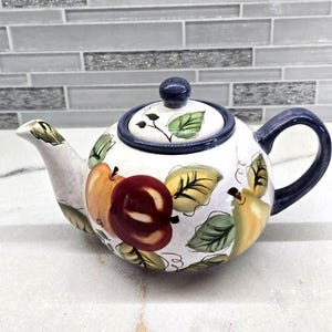 May include: A white ceramic teapot featuring a hand-painted fruit motif with red apples, yellow pears, and green leaves. The teapot includes a matching lid with a dark blue knob, a curved spout, and a dark blue handle, all with a glossy finish.