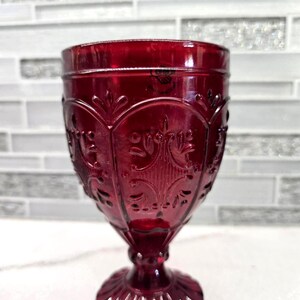 May include: A ruby red goblet with an ornate, embossed design. The glass features a detailed pattern of swirling lines and floral motifs. The goblet has a round base and a stem that leads to the bowl. The glass is made of a transparent material.