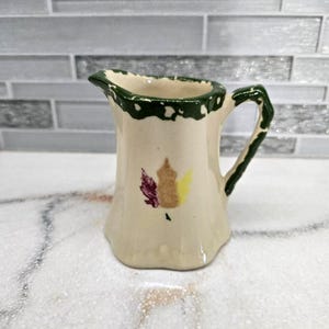 May include: A small, off-white ceramic pitcher with a green rim and handle. The pitcher features a leaf design in shades of purple, yellow, and brown. The pitcher is likely used for serving cream or syrup.