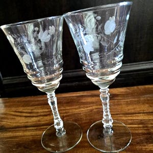 May include: Two clear, stemmed wine glasses with etched floral designs. The glasses have a flared bowl, a decorative stem, and a round base. The glasses are positioned on a wooden surface.