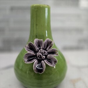 May include: A small, green ceramic vase with a glossy finish. A decorative, dark gray flower is affixed to the front. The vase has a bulbous base and a narrow neck, suitable for displaying small floral arrangements.