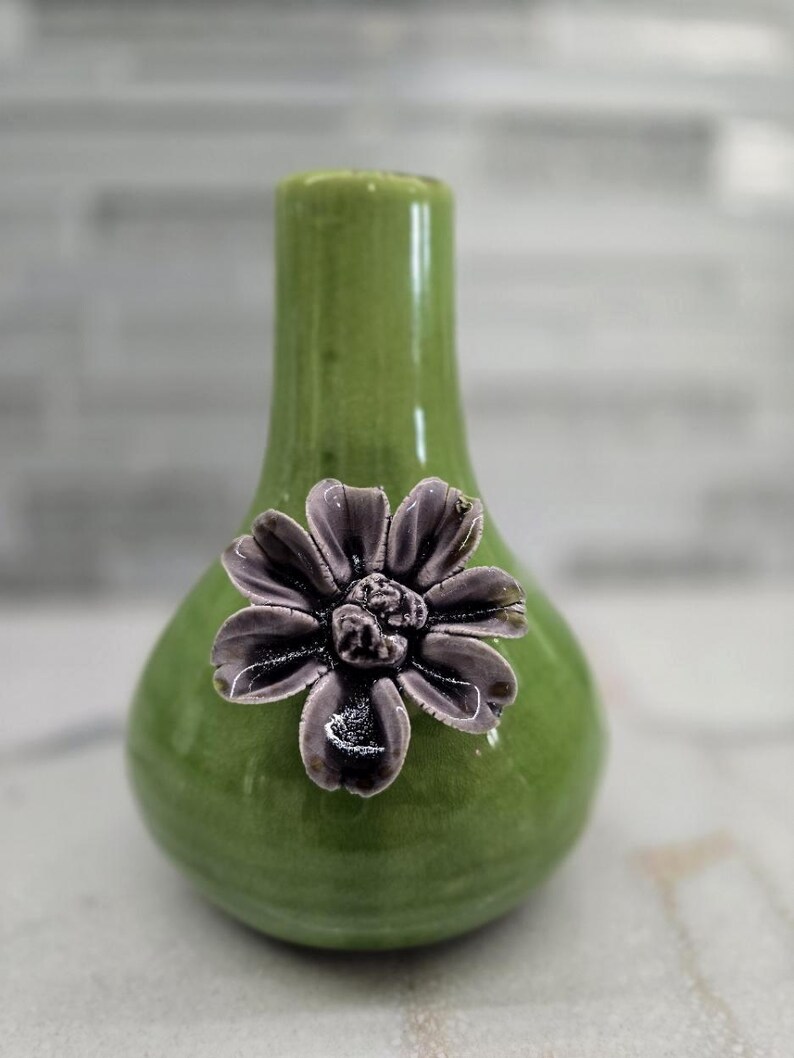 May include: A small, green ceramic vase with a glossy finish. A decorative, dark gray flower is affixed to the front. The vase has a bulbous base and a narrow neck, suitable for displaying small floral arrangements.