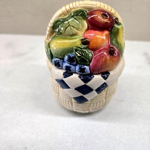 May include: A ceramic salt and pepper shaker shaped like a woven basket filled with assorted fruits including apples, pears, and blueberries. The basket features a cream-colored exterior with a blue and white checkered cloth draped over the front.
