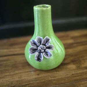 May include: A small, green ceramic vase with a bulbous body and a narrow neck. A three-dimensional, gray and purple flower is affixed to the front. The vase is glossy and sits on a wooden surface.