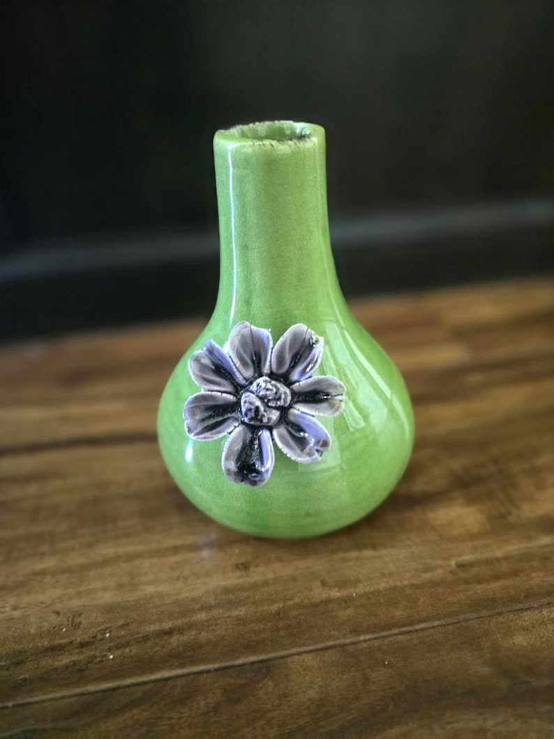 May include: A small, green ceramic vase with a bulbous body and a narrow neck. A three-dimensional, gray and purple flower is affixed to the front. The vase is glossy and sits on a wooden surface.