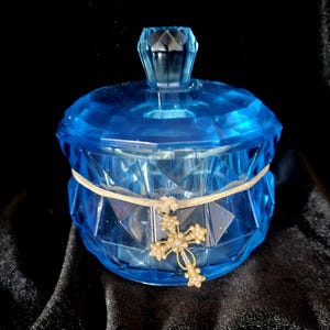 May include: A faceted, blue glass jar with a matching lid. A cream-colored ribbon is wrapped around the jar, and a gold-colored cross pendant with small, clear stones hangs from the ribbon. The jar sits on a black surface.