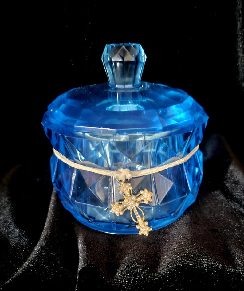 May include: A faceted, blue glass jar with a matching lid. A cream-colored ribbon is wrapped around the jar, and a gold-colored cross pendant with small, clear stones hangs from the ribbon. The jar sits on a black surface.