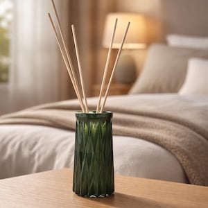 May include: A green glass diffuser bottle with a geometric pattern, holding several wooden reeds. The bottle sits on a wooden nightstand, with a bed and lamp in the background. This item is perfect for home fragrance.