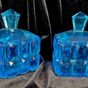 May include: Two matching, faceted, blue glass trinket boxes with geometric lids. The boxes have a square shape with multiple facets, creating a sparkling effect. The lids are also faceted and have a diamond-like shape.